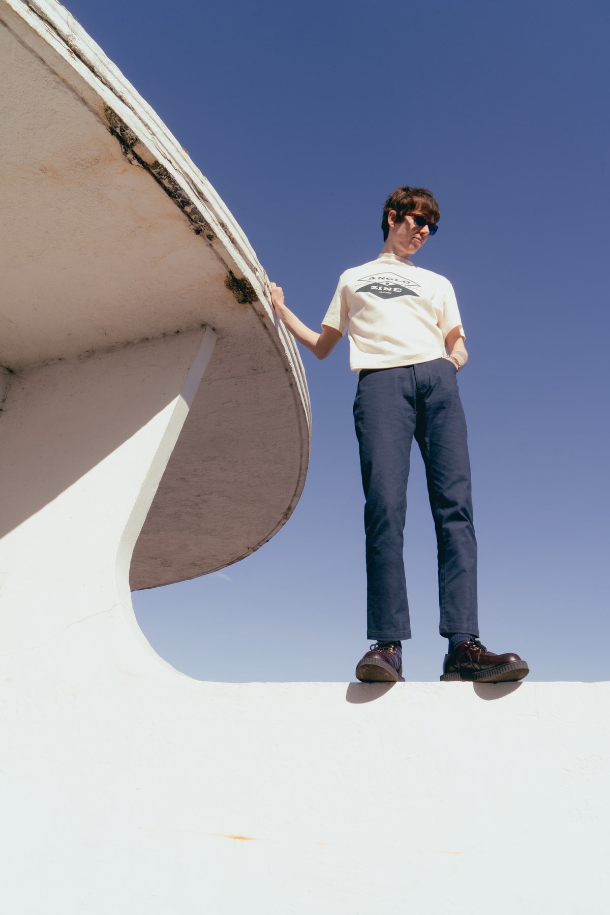 Model wearing an Anglozine Beppo print T-shirt and navy trousers beside curved white architecture.